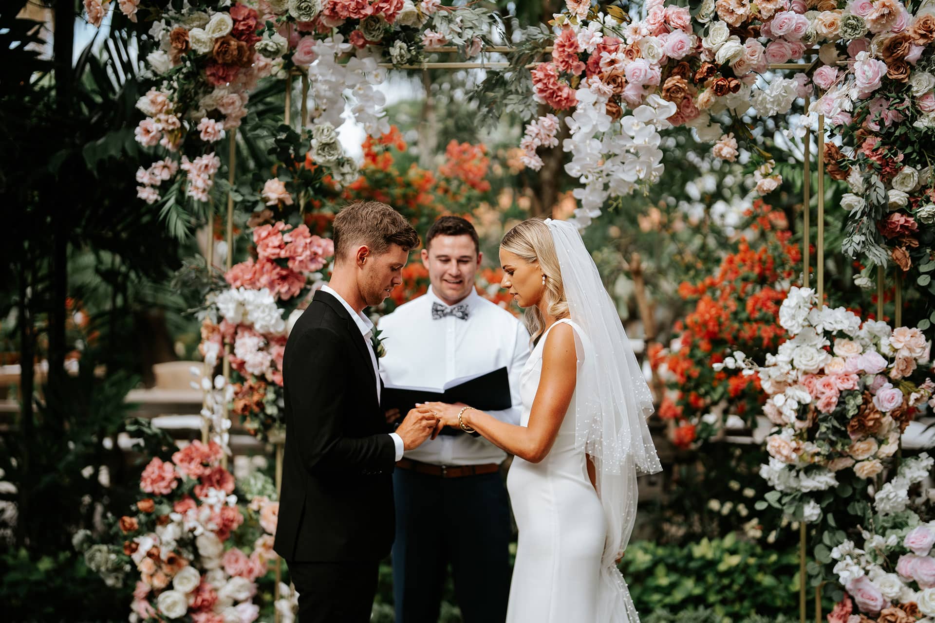 A bride and groom hold a wedding ceremony under a flower arch with a priest