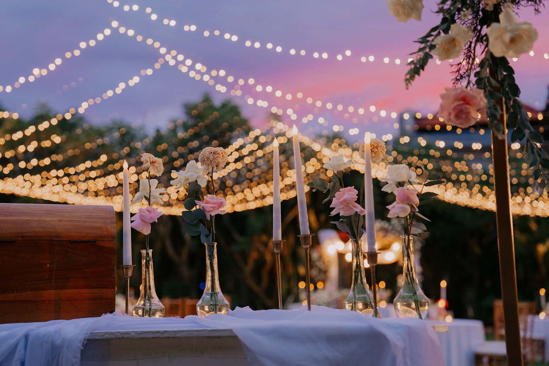 An outdoor dining table decorated with candles, flower vases and hanging lights in the background at sunset