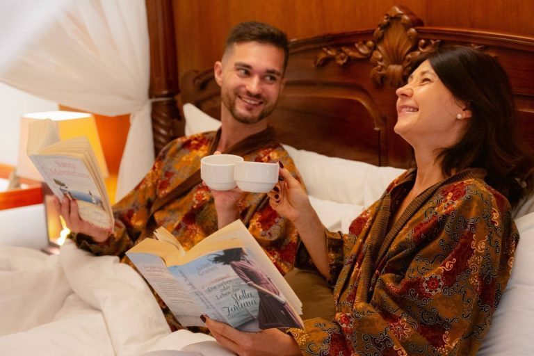 A relaxed couple enjoying coffee and reading in bed, experiencing a premium long-term holiday lifestyle at Astagina Resort Villa and Spa.