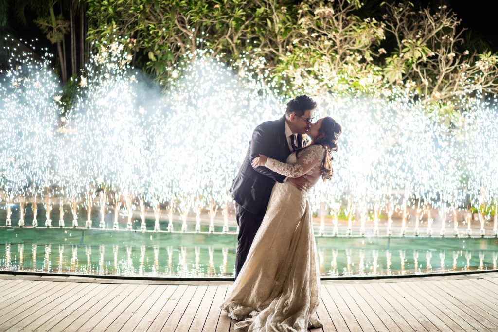 Bride and groom sharing first kiss in front of cold spark pyrotechnics display at outdoor evening wedding reception at Astagina Resort Legian Bali
