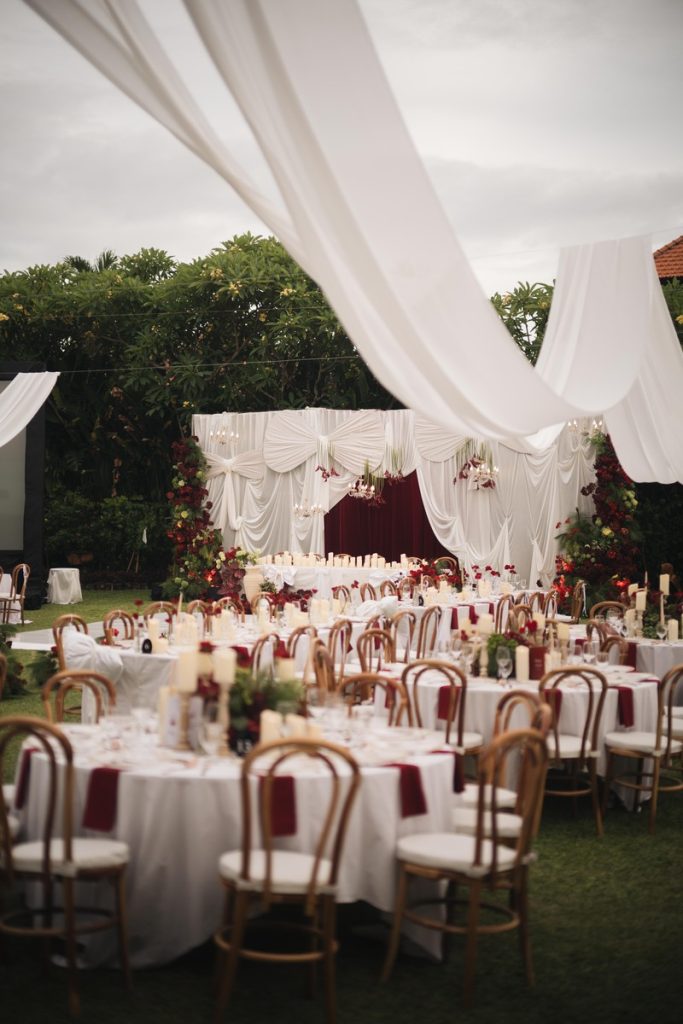 Grand wedding reception setup at Astagina Resort Bali featuring elegant white draping, red floral decor, and round guest tables