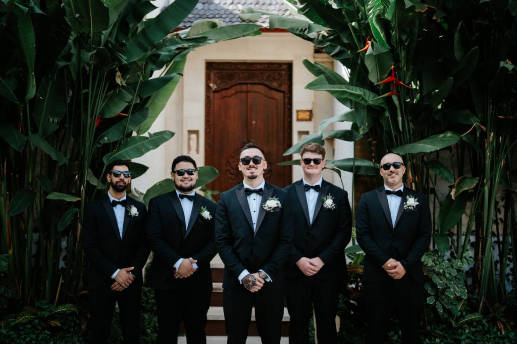 Groom and groomsmen in black tuxedos under tropical palms, Astagina Resort wedding Legian